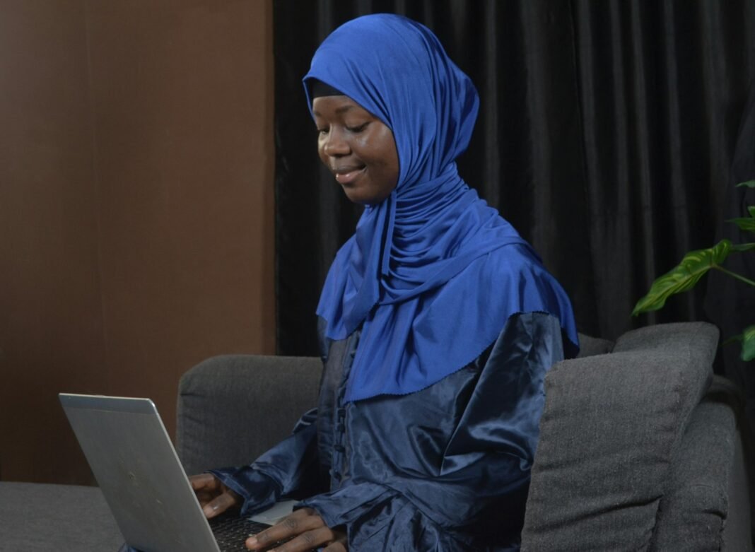 Rent Is Due in 12 Months. Here’s How to Be Ready Without Borrowing Young woman in a blue headscarf sitting on a couch with a laptop, planning her rent savings