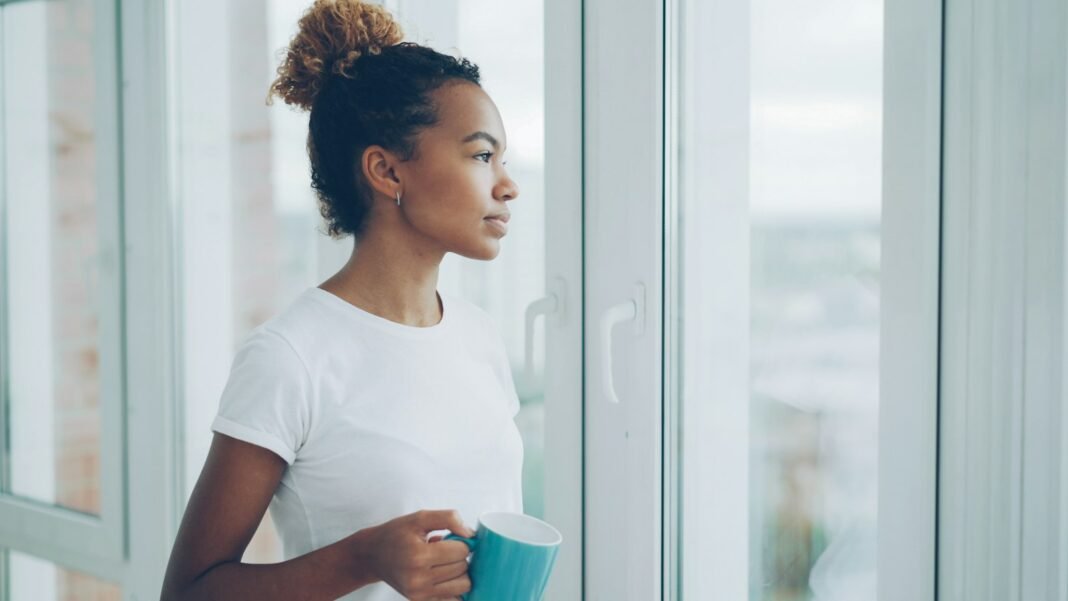 New City, Full Calendar, Still Lonely? You’re Not Alone Young Black woman holding a mug and looking out of an apartment window, reflecting on loneliness in the city