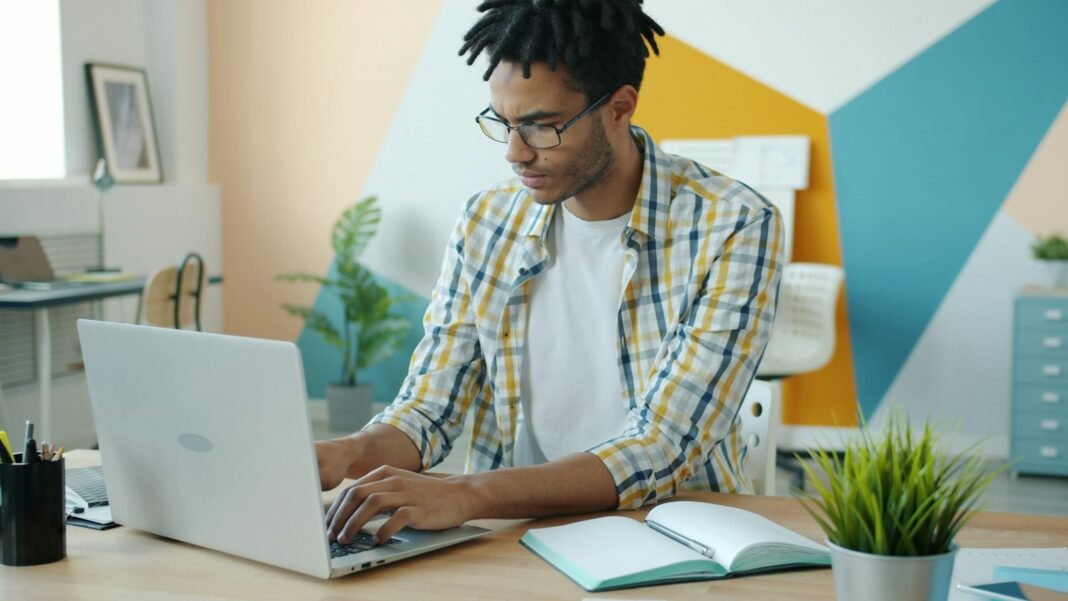 Stop Asking AI Vague Questions: Use These Work Prompts Instead Young Black man with dreadlocks typing on a laptop at his desk, writing AI prompts for work