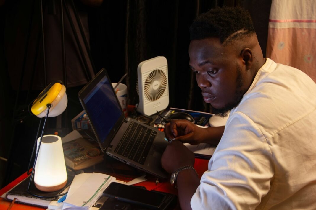 Young Nigerian man studying on a laptop at night under desk lamps after work