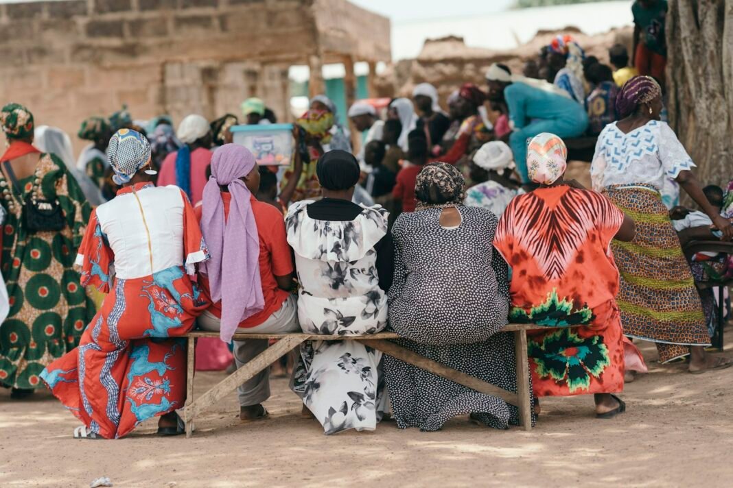 Ajo, Esusu or Cooperative? How to Save Together Without Losing Money Women in colourful clothes seated on benches at an outdoor community meeting, like members of a savings group