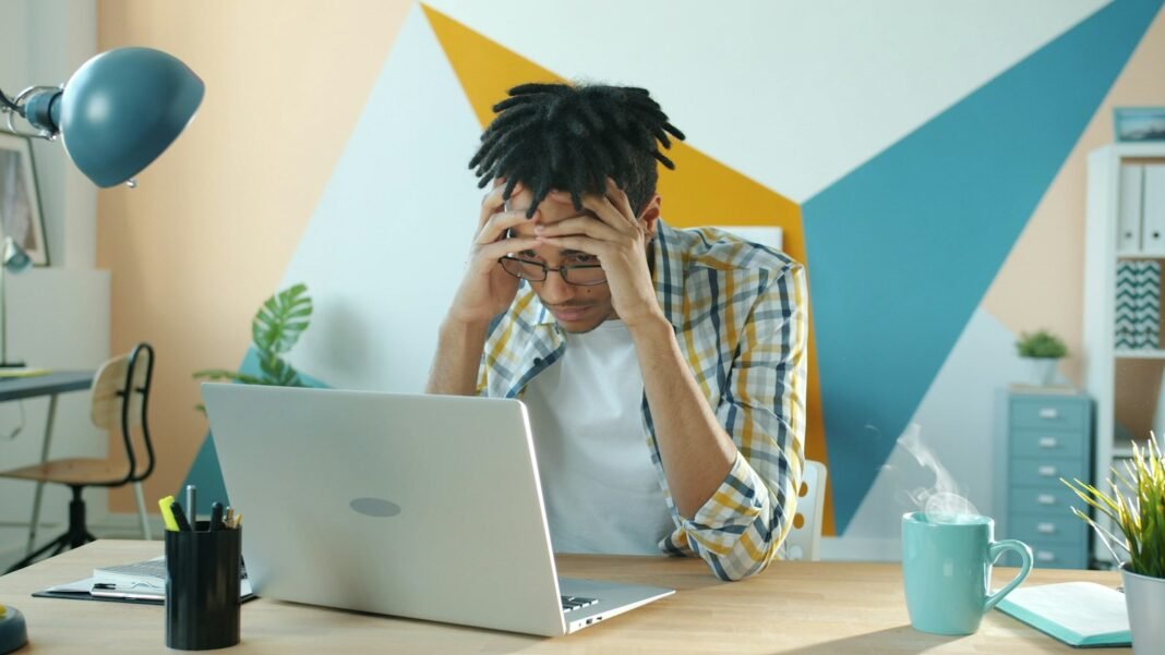 Money Worries Keeping You Up at Night? How Financial Stress Affects Your Mind Young Black man holding his head in frustration at a desk with a laptop, showing money stress